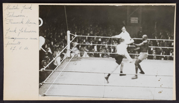 Schwarz-weiß-Foto von zwei Boxern, Jack Johnson und Frank Morgan, in einem Boxring mit Zuschauern im Hintergrund.