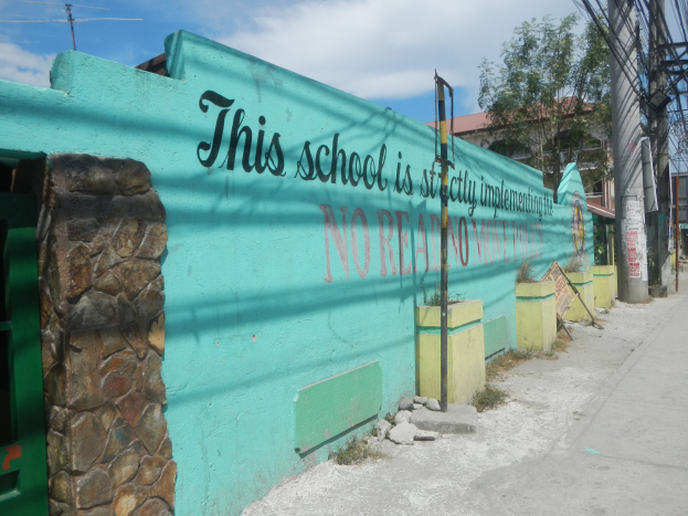 Grüne Wand mit Graffiti, das 'Diese Schule ist keine Stadt' lautet, umgeben von Gebäuden, Bäumen, Strommasten, Drähten und anderen städtischen Elementen unter einem bewölkten Himmel.