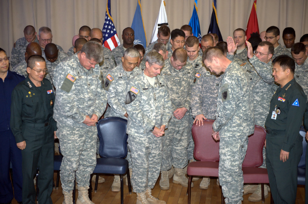 Eine Gruppe von Menschen, hauptsächlich in militärischer Kleidung, steht zwischen roten und blauen Stühlen mit Flaggen im Hintergrund.