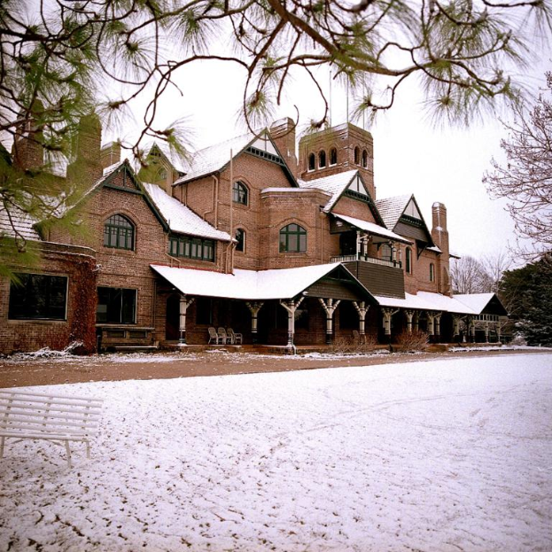 Ein Gebäude mit Fenstern und Säulen, Stühle davor, eine Bank und ein Zweig auf der rechten Seite.