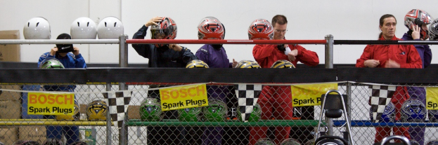 Ein Maschendrahtzaun mit Plakaten und Fahnen daran, mit einigen Menschen im Hintergrund, einige tragen Helme, und zusätzliche Helme auf einem Ständer, alles vor einer Wand.