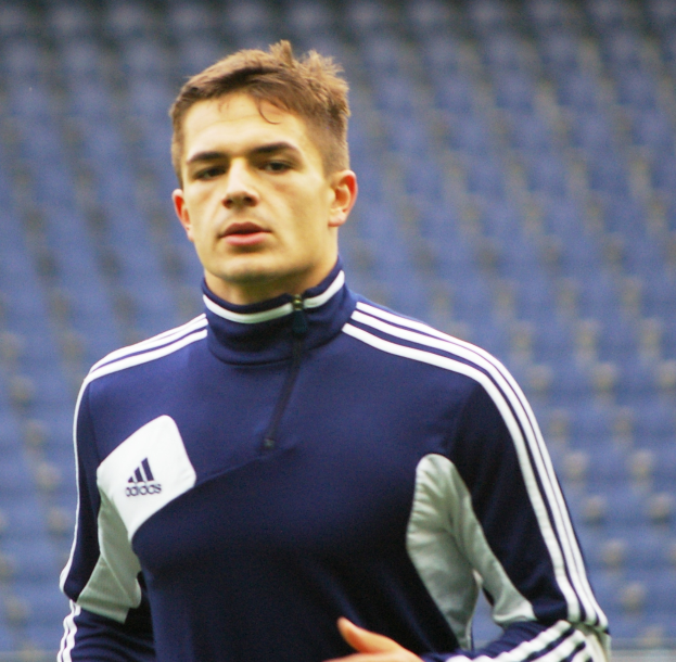 Mann in blauem Jackett auf Fußballfeld mit ausgestreckten Armen und entschlossener Miene, mit unscharfem Hintergrund.