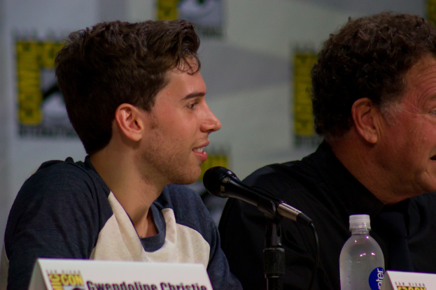 Ein Mann lächelt vor einem Mikrofon, mit zwei Namensschildern und einer Flasche in der Nähe und einem Banner im Hintergrund.