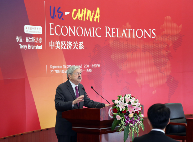 Ein Mann in einem schwarzen Blazer, weißem Hemd und lila Krawatte steht an einem Podium mit zwei Mikrofonen und einem Blumenstrauß, spricht vor einer roten Fahne mit der Aufschrift 'US-China Economic Relations.'