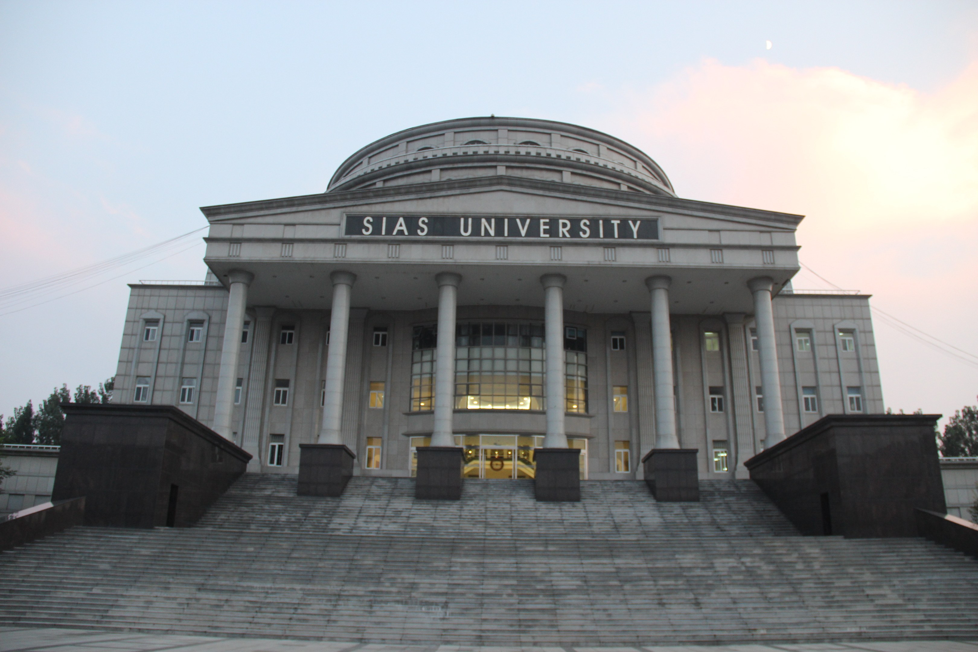 Large university building with pillars, windows, and steps, surrounded by trees under a clear blue sky, with the university's name visible on the facade.
