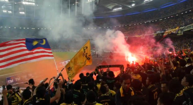 Large crowd in a stadium with flares, flags, and smoke during a live streaming event labeled "Malaysia vs Malaysia Live Streaming".