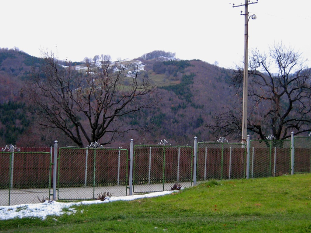 Eine grasbewachsene Landschaft mit einem Zaun und einem Pfahl im Vordergrund, Bäume verstreut und einige Häuser im Hintergrund auf einem Hügel sichtbar.