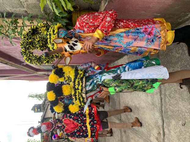 Eine Gruppe von Menschen in farbenfrohen Kostümen steht zusammen vor einem Gebäude, mit verschiedenen Gegenständen in der Hand, vor einem Hintergrund aus Bäumen, Gebäuden und einem klaren blauen Himmel während eines Karnevalsumzugs.
