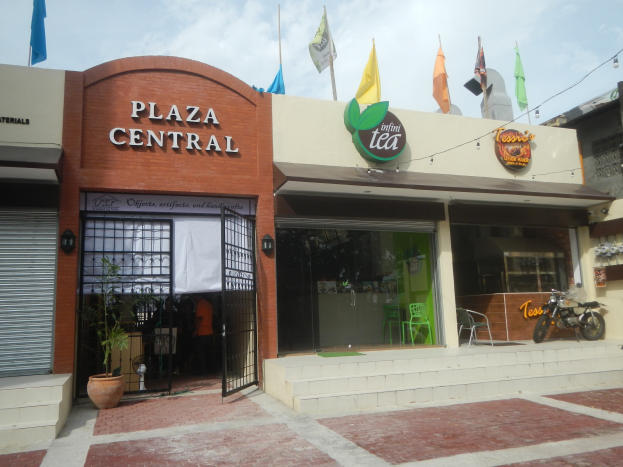 A central plaza in Cebu City, Philippines, with buildings, motorbikes, outdoor seating, plants, flags, signage, overhead electric cables, and a cloudy sky.