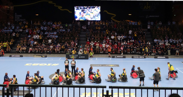 Eine Gruppe von Athleten im Rollstuhl auf einem Platz bei den Invictus Games Düsseldorf 2020, umgeben von einer Menge, mit Treppen zu einem Bildschirm und Informationsplakaten im Hintergrund.