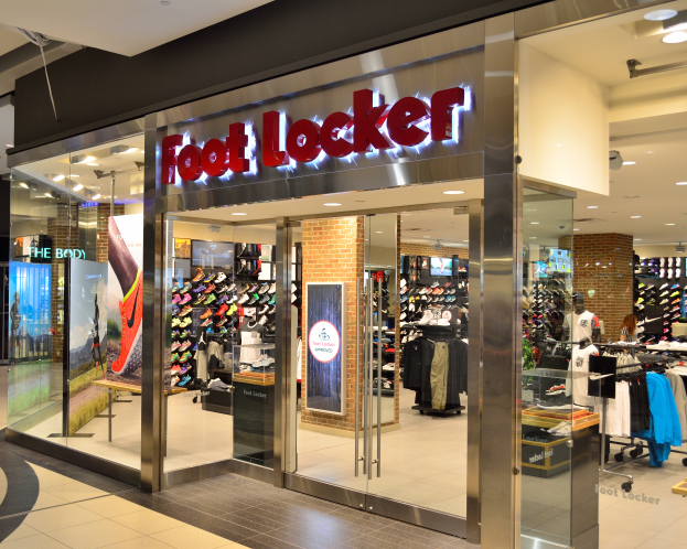 Ein Foot Locker Geschäft in einem Einkaufszentrum, mit Glaswänden und -türen, Kleidung an Bügeln, bekleidete Schaufensterpuppen, Texttafeln, Deckenleuchten und verschiedene andere Gegenstände.