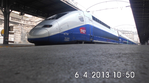 Ein Zug ist in der Mitte des Bildes mit einer Plattform und Text unten, Gebäuden und einem Dach links und Gebäuden, Dächern, Kabeln und Himmel oben.