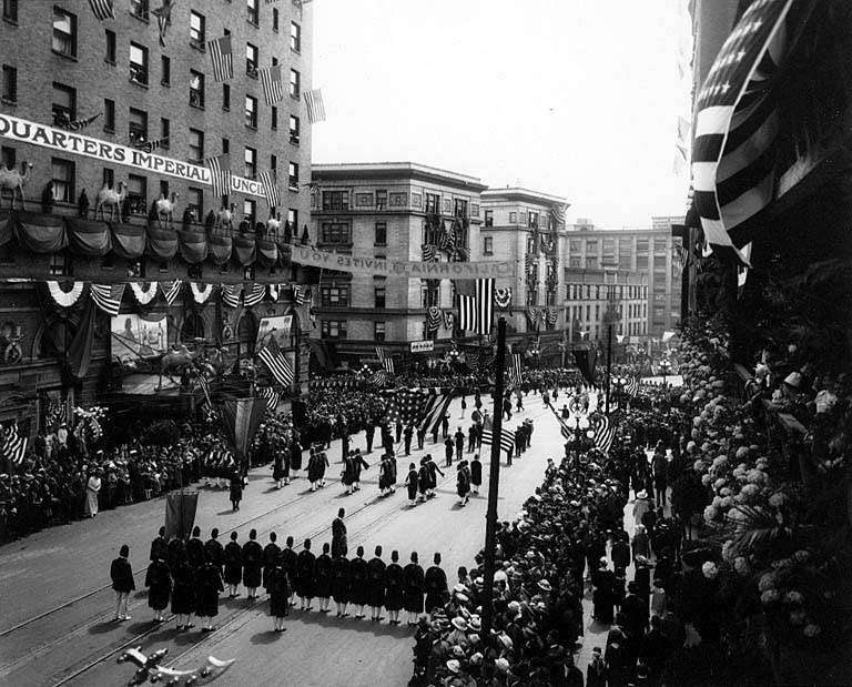 A large crowd of people walks down a New York City street during a Fourth of July parade, many wearing caps and holding flags, with others lining the sides; flags on poles and trees line the street, buildings with windows and a distant sky are visible, and text is written on one building.