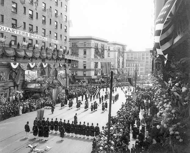 A large crowd of people walks down a New York City street during a Fourth of July parade, many wearing caps and holding flags, with others lining the sides; flags on poles and trees line the street, buildings with windows and a distant sky are visible, and text is written on one building.