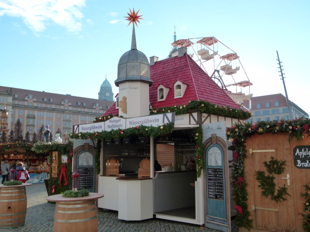 Kleines Gebäude mit rotem Dach und einem Riesenrad davor, umgeben von Festdekorationen und Menschen, mit Gebäuden, Bäumen und einem bewölkten Himmel im Hintergrund auf einem Oktoberfest in München, Deutschland.