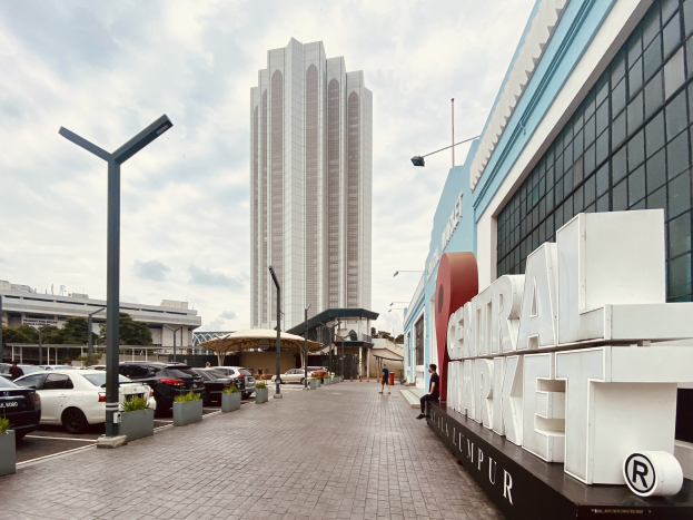 A bustling central market in Kuala Lumpur, Malaysia, featuring tall buildings, street poles, street lights, motor vehicles, pedestrians on the footpath, name boards, trees, and a cloudy sky.