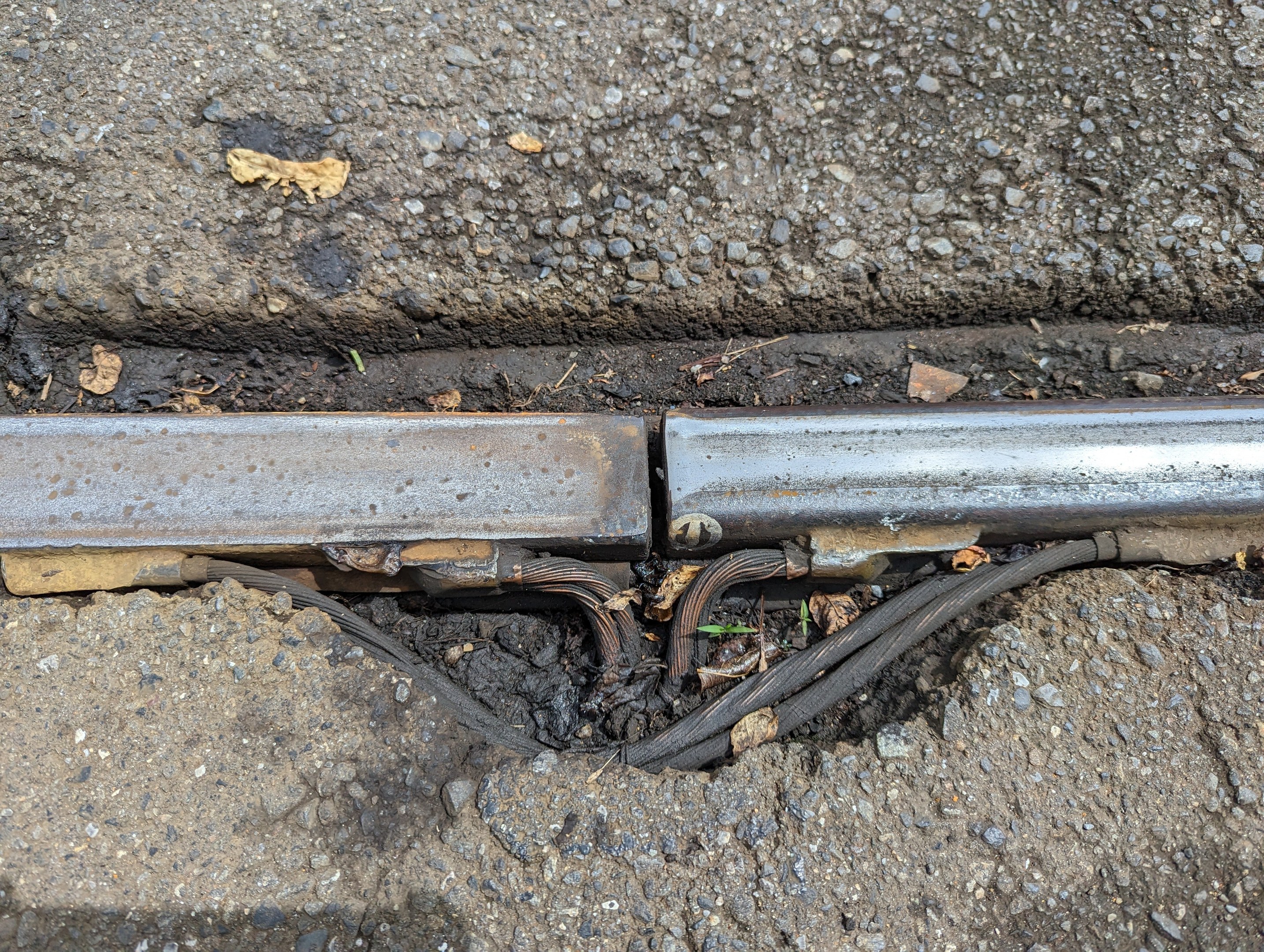 Nahaufnahme eines Metallkanalrohrs an der Seite einer Straße mit verstreuten Drähten und trockenen Blättern, das mit dem Boden verbunden ist.