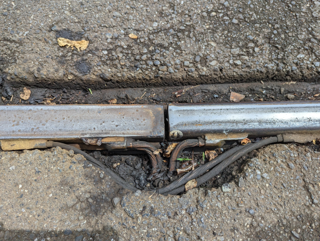 Nahaufnahme eines Metallkanalrohrs an der Seite einer Straße mit verstreuten Drähten und trockenen Blättern, das mit dem Boden verbunden ist.