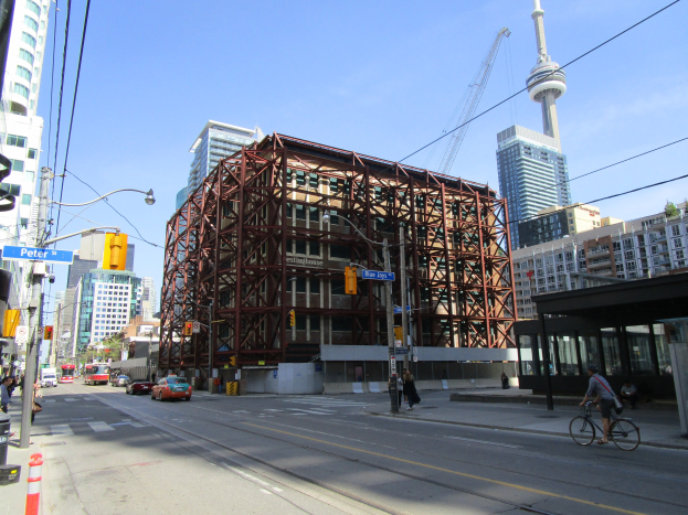 Eine belebte Stadtstraße mit einem großen Baugerüst im Hintergrund, das Fahrzeuge, einen Radfahrer, Fußgänger, Ampeln, Pfosten, Schilder, einen Kran und den Himmel zeigt.