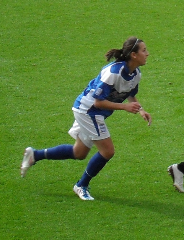 Eine Frau in einer blauen und weißen Uniform rennt auf einem Fußballfeld und trägt Fußballschuhe, wobei ein Personenschlag auf der rechten Seite des Bildes zu sehen ist.