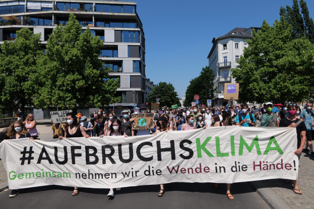 Eine Gruppe von Menschen mit Masken hält ein Banner mit der Aufschrift 'Aufbruchsklima' vor einem Gebäude mit Bäumen und einem klaren Himmel.