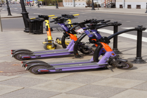 Eine Reihe von E-Scootern, die an einer Straße geparkt sind, mit einem Müllcontainer im Vordergrund und Gebäuden, Bäumen und Fahrzeugen im Hintergrund.