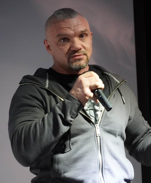 Comedian in einem schwarzen Anzug und weißem Hemd, der ein Mikrofon hält und selbstbewusst vor einer Wand steht.