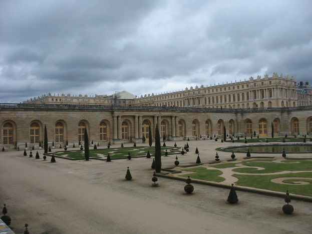 Ein prächtiger Palast mit kreisförmigen Pflanzen im Vordergrund und einem bewölkten Himmel darüber.