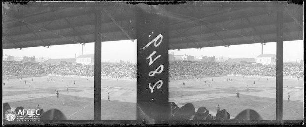 Ein Schwarz-Weiß-Foto eines Fußballspiels in einem Stadion, mit Spielern auf dem Boden und Zuschauern in den Rängen, vor Gebäuden, Bäumen und einem klaren Himmel, mit dem Text "Piazza della Repubblica, Rom, Italien" in der linken unteren Ecke.