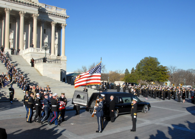 Funeral procession with people in caps and flags in front of the U.S. Capitol Building, vehicles on the road, steps with railings, light poles, trees, and a sky in the background.