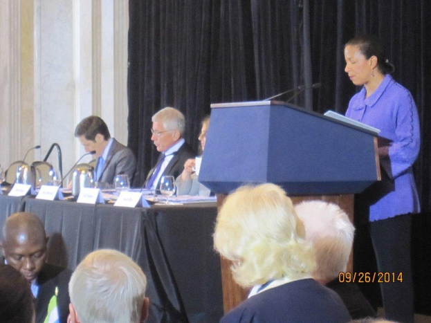 Eine Frau steht an einem Tisch und spricht während einer Konferenz, mit drei sitzenden Männern neben ihr an einem anderen Tisch mit Gläsern, Büchern und Namensschildern, während einige Menschen vor den Tischen arbeiten und ein Vorhang und eine Wand im Hintergrund zu sehen sind.