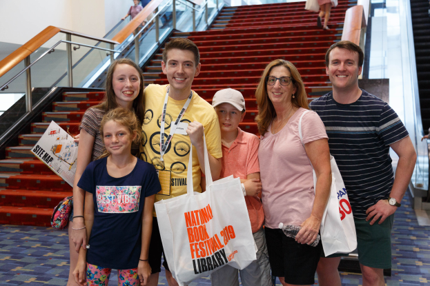 Eine Gruppe von Menschen, die zusammenstehen, lächeln und Taschen halten, vor einer Rolltreppe auf der Nationalen Buchmesse 2019, mit Treppen, Geländern und einer Wand im Hintergrund.