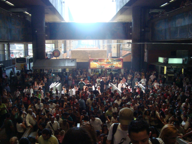Eine Menschenmenge, die vor Marktständen steht, mit fahrenden Fahrzeugen im Hintergrund und Säulen an der Wand.