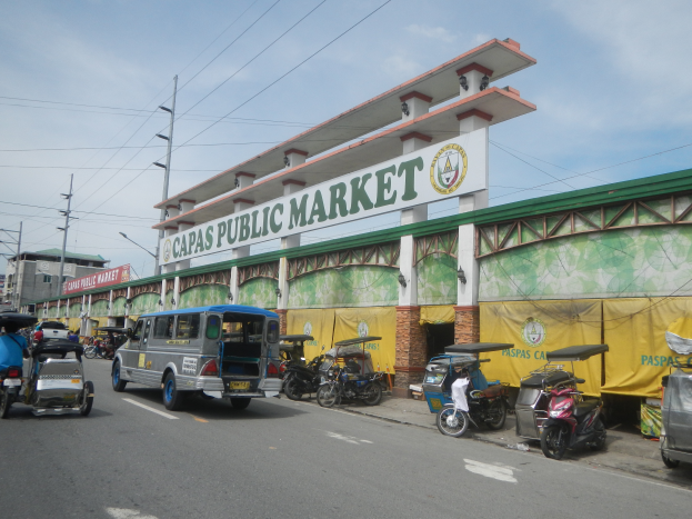 Eine belebte Straße mit Autos, Motorradfahrern und Rikschas, die an dem Gebäude des Capas Public Market vorbei fahren, mit Strommasten, Laternen und bewölktem Himmel im Hintergrund.