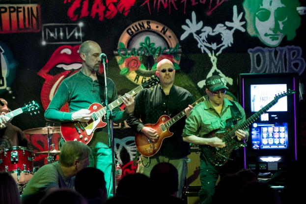 Drei Männer auf einer Bühne bei einem Konzert, mit einer Menge darunter und einer bemalten Wand im Hintergrund.