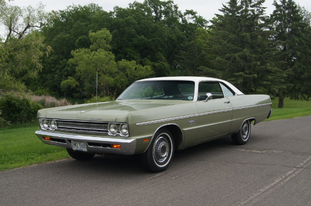 Ein grünes Oldtimer-Auto fährt auf einer Straße mit Bäumen im Hintergrund und einem klaren Himmel darüber.