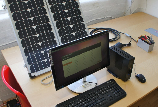 Ein Computersystem mit Tastatur, Maus und CPU auf einem Tisch, neben einer Solarpaneel, Batterie, Chips und Kabeln, mit einem roten Stuhl und weißer Wand im Hintergrund.