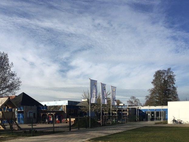 Großes Gebäude mit einem großen Rasenbereich, Metallzaun, Weg, Bäumen, Pfählen, Bannern, einem abgestellten Fahrrad, nahen Häusern und einem bewölkten Himmel, identifiziert als der neue Spielplatz der Schule.