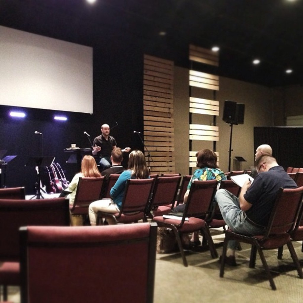 In einer Halle sitzen mehrere Frauen auf roten Ledersesseln, während ein Mann in einem schwarzen Hemd vor ihnen spricht. Hinter ihm ist ein weißer Projektionsschirm, eine hölzerne Dekoration an der Wand und ein Lautsprecher auf einem Ständer. Die Decke ist schwarz mit Scheinwerfern.