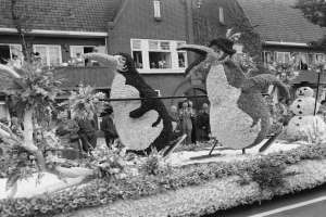 Schwarzes und weißes Foto eines Paradewagens, der mit Blumen und Pinguinfiguren geschmückt ist, mit Menschen auf der Straße und Gebäuden im Hintergrund.